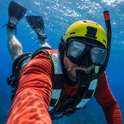 A person snorkeling underwater, wearing a yellow helmet, black mask and snorkel, a red long-sleeve shirt, and a black and white life vest, with blue ocean water in the background.